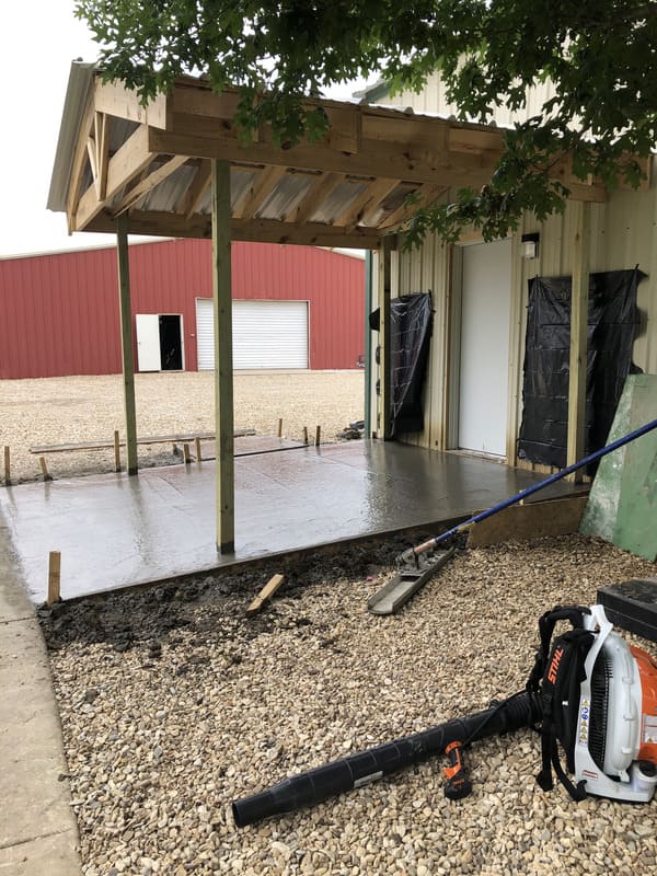 Freshly poured concrete slab beneath a wooden awning outside a beige metal building, with a Stihl backpack leaf blower and gravel in the foreground.