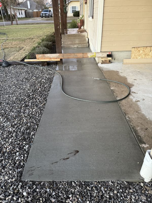 Newly poured concrete sidewalk next to a house, bordered by gravel and a garden hose lying on it.