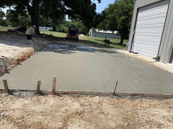 Worker smoothing freshly poured concrete slab outside a metal building with trees and lawn in the background.
