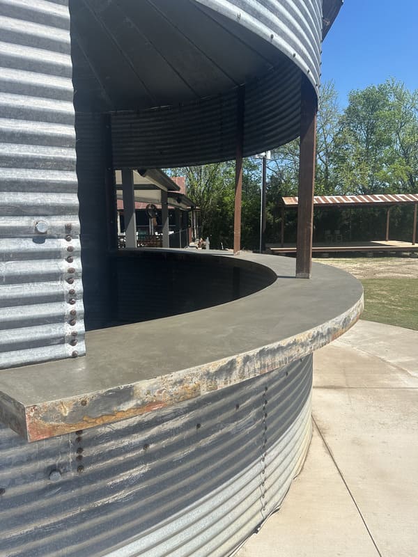 Curved concrete countertop attached to a corrugated metal structure under a clear blue sky with trees and outdoor shaded seating in the background.