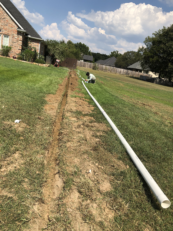 Person installing a long white drainage pipe next to a narrow trench dug in a grassy backyard beside a brick house.