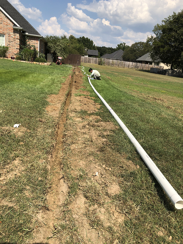 Person kneeling on a lawn installing a long white drainage pipe along a freshly dug trench beside a red brick house under a partly cloudy sky.