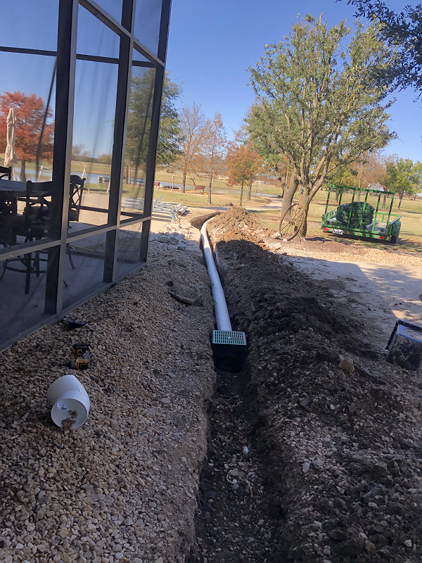 Outdoor drainage installation beside a house with gravel and soil, featuring a white pipe connected to a green drainage grate under a tree.