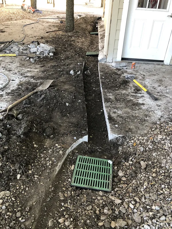 Trench dug beside a house with green drainage grates installed and construction tools nearby on a dirt and gravel surface.