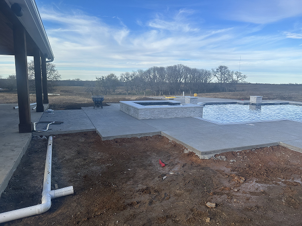 Partially constructed outdoor pool area with concrete decking, a hot tub, exposed earth, and a white drainage pipe under a cloudy sky.