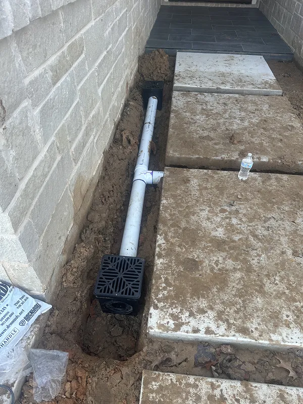 Outdoor drainage system with white PVC pipes and black grates installed alongside a stone wall and next to large concrete slabs.