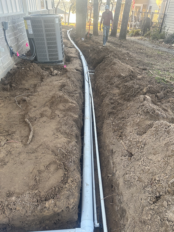 Trenched ground alongside a house with large gray drainage pipes laid in the trench and a man walking nearby.