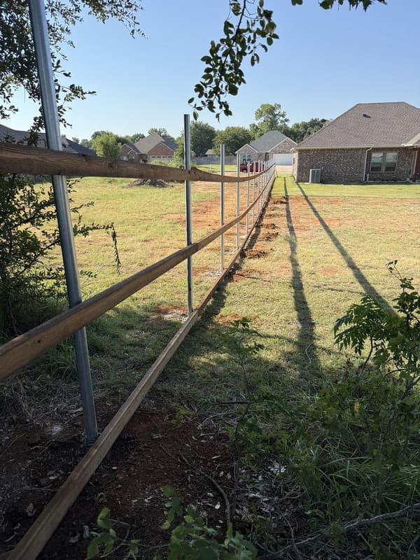 Partially constructed wooden fence with metal posts stretching across a grassy yard toward suburban houses under a clear blue sky.