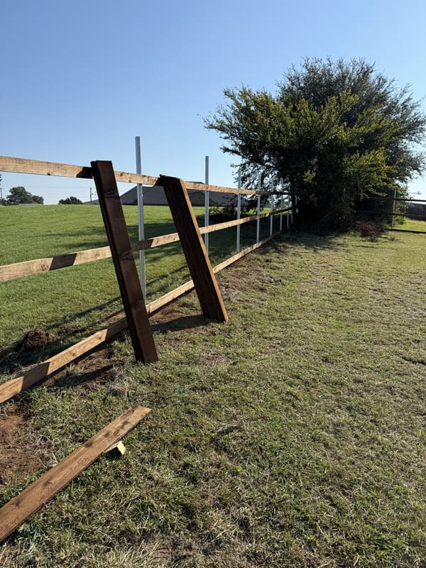Partially constructed wooden fence along a grassy field with two upright fence panels leaning against it and a green bush in the background.