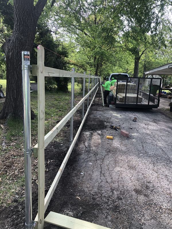 Wooden fence frame being constructed along a driveway with workers and a trailer loaded with materials in the background.
