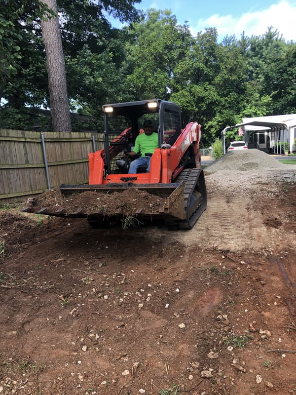 Man operating an orange compact track loader moving dirt on a residential driveway with gravel piles in the background.