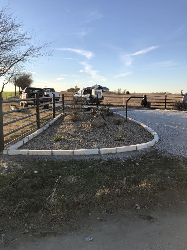 Gravel driveway bordered by white concrete blocks with a metal fence on the left and two trucks parked near a rural house under a blue sky.
