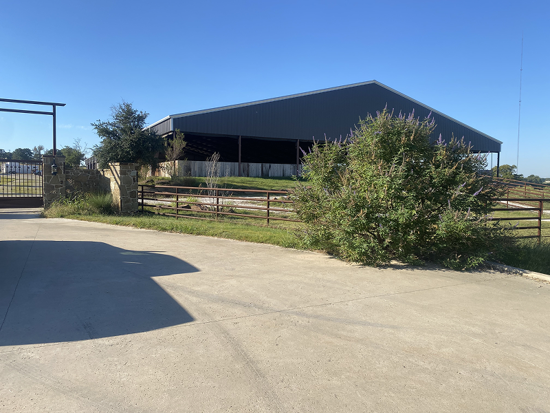 Large open metal barn with fenced grassy area and a bush in front, under a clear blue sky.