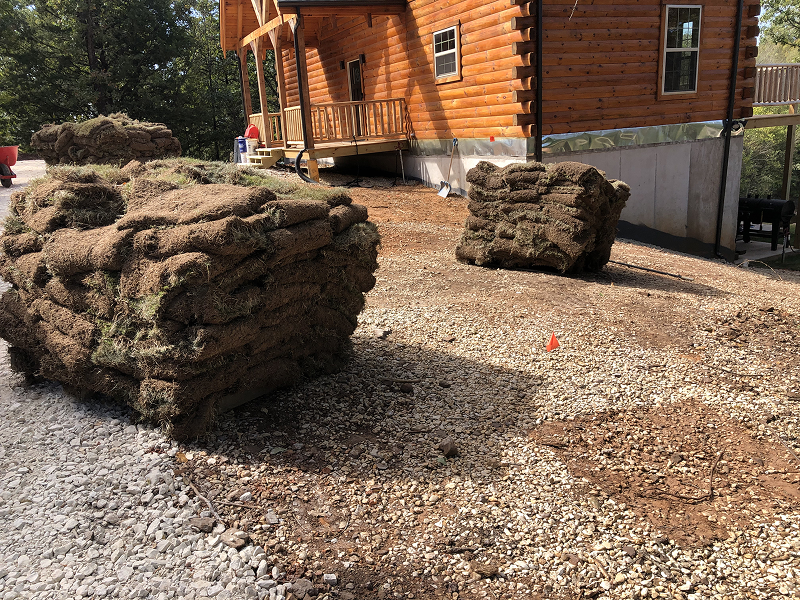 Stacks of rolled sod placed on gravel near a wooden cabin under construction.