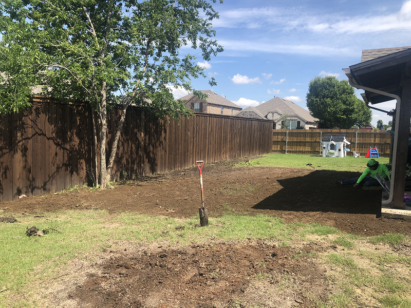 Backyard with soil preparation for sod installation, showing a shovel stuck in the ground and a person working near a house under a sunny sky.