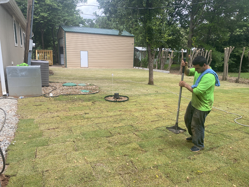 Worker installing sod in a yard beside a house, using a long-handled tool to press the grass into the soil.