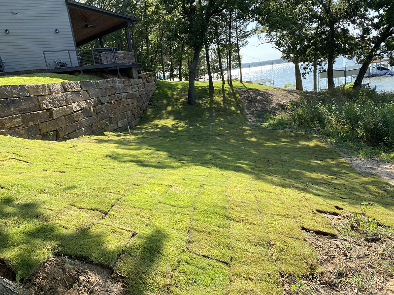 Newly laid sod on a sloped lawn next to a stone retaining wall and a house, with a view of a lake and docks beyond trees.