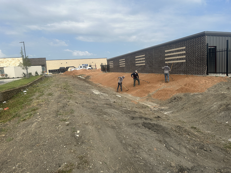 Three construction workers raking red soil next to a modern brick and metal building under a partly cloudy sky.