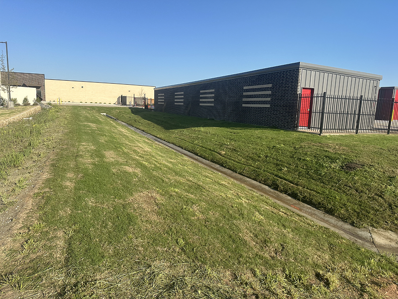 Sloped grassy area with a narrow concrete drainage channel running alongside a dark brick and metal building with red doors under a clear blue sky.
