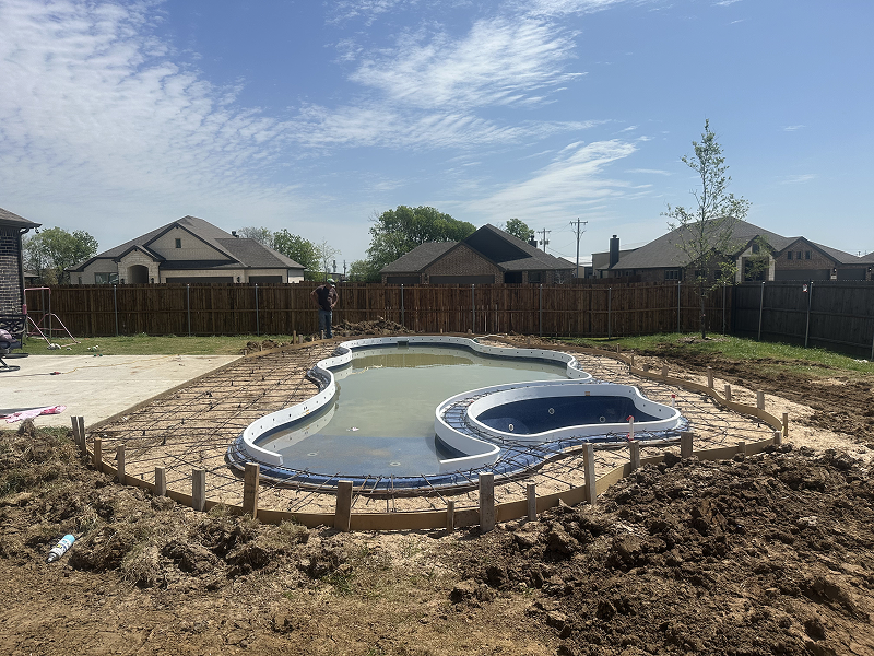 Partially constructed backyard swimming pool with water inside, surrounded by wooden forms and metal rebar for concrete pouring under a blue sky.