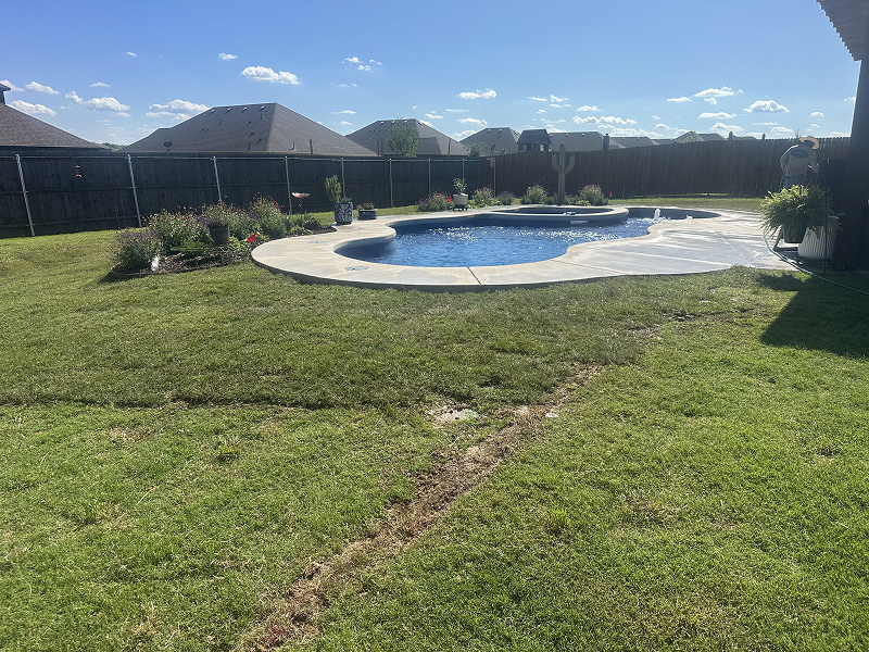 Backyard with a kidney-shaped swimming pool surrounded by a concrete deck and grassy lawn under a blue sky with some clouds.