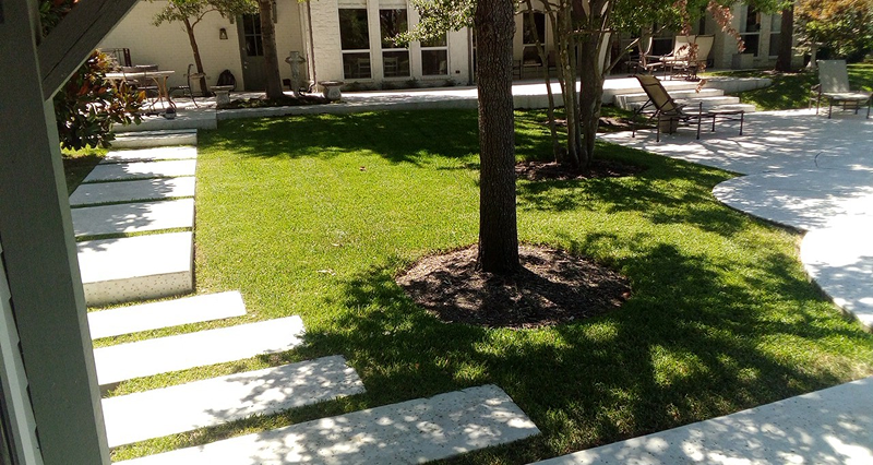 Backyard with freshly cut green grass, a central tree with mulch around the base, stepping stones on the left, and patio furniture on the right side.
