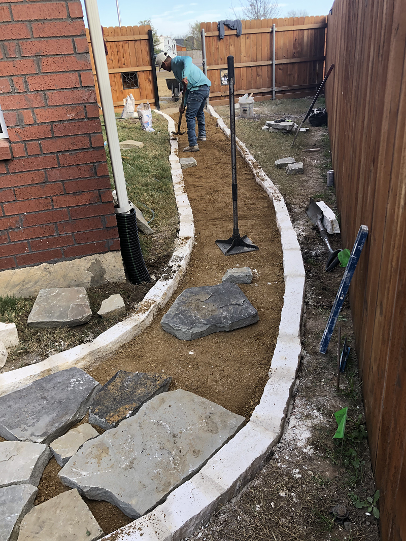 Person in blue shirt and cap working on installing stone walkway bordered by white bricks between a brick house and wooden fence.
