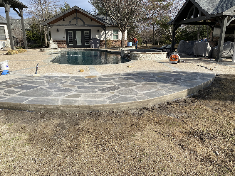 Curved stone patio in front of a backyard swimming pool with a pool house and pergola nearby.