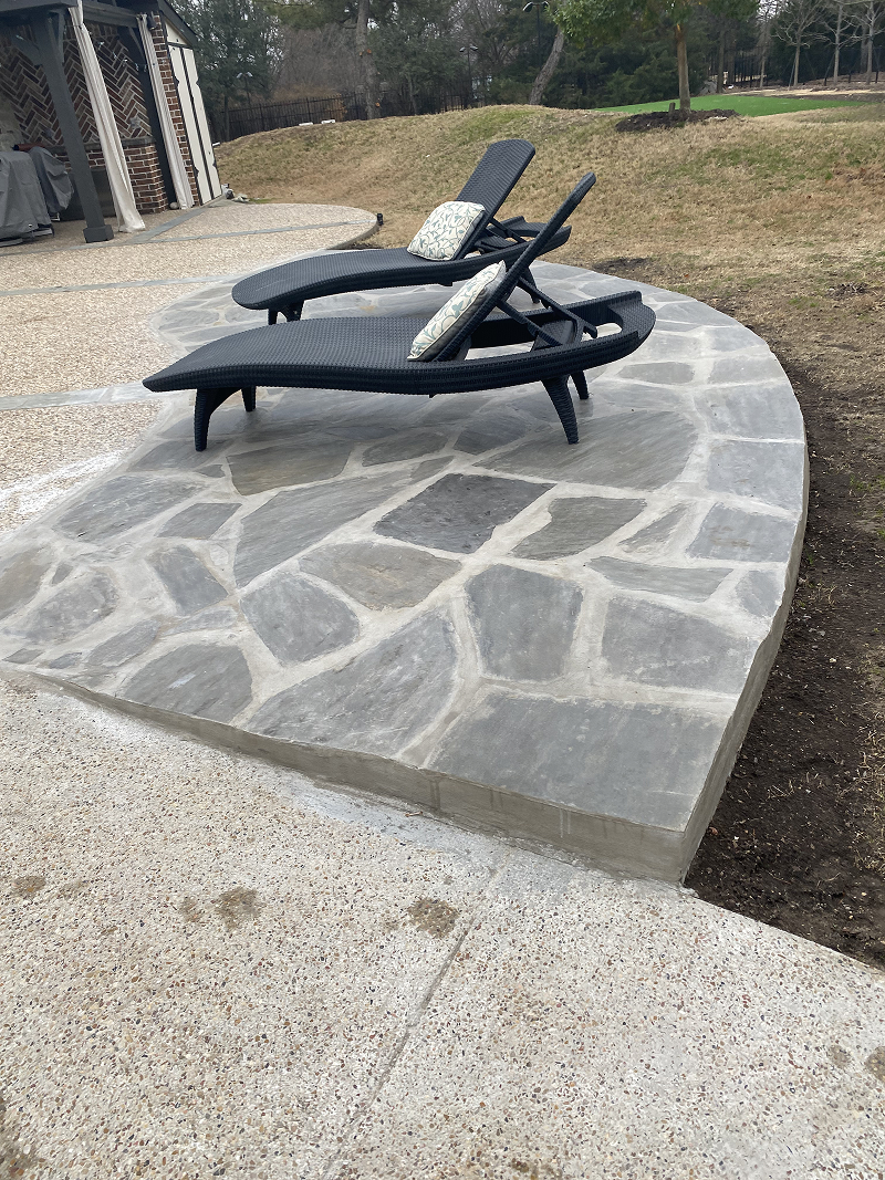 Two black woven lounge chairs with patterned cushions on a curved stone patio surrounded by grass and a concrete surface.