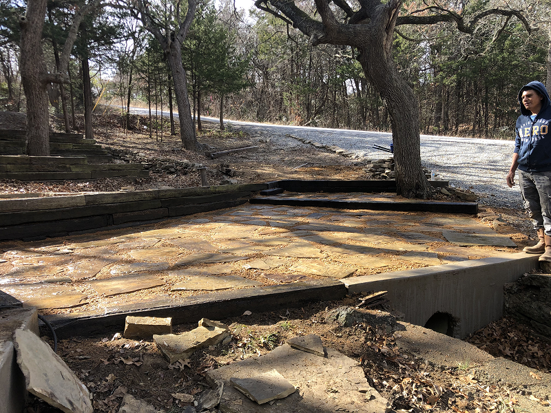 Stone walkway with irregular flagstones and a tree in the middle, next to a gravel road and surrounded by forest.