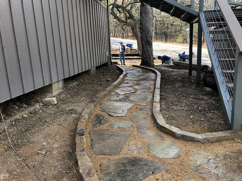 Stone walkway bordered by bricks running alongside a building and beneath a metal staircase with two workers in the background.