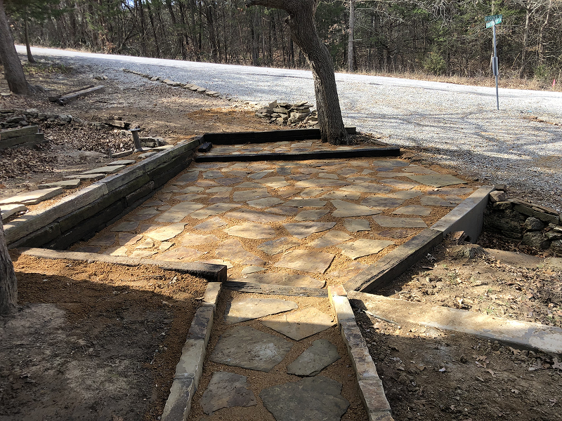 Stone walkway and patio area with a tree in the center, bordered by wooden beams and surrounded by dirt and gravel road in a wooded area.