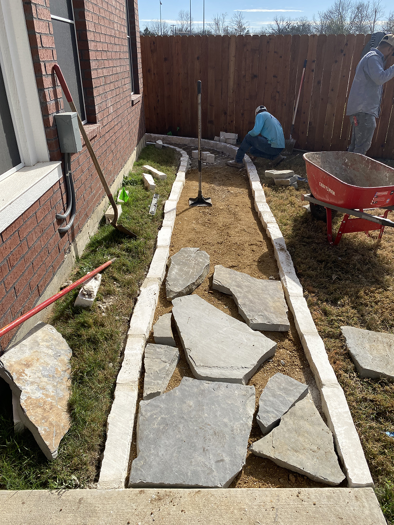 Construction of a stone walkway with large irregular flagstones laid on dirt path bordered by small rectangular stones, with two workers in the background and a red wheelbarrow nearby.