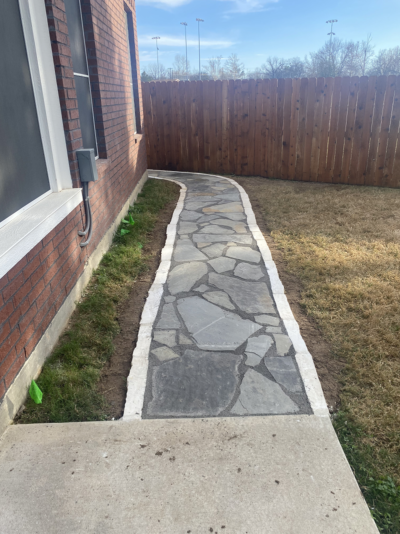 Stone walkway with white border running alongside a brick house wall, leading to a wooden fence.