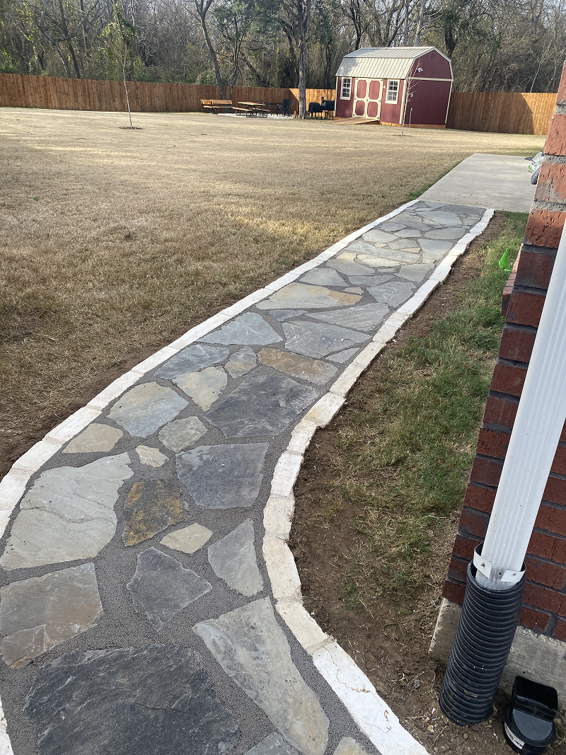 Stone walkway bordered with white edging leading to a red wooden shed in a fenced backyard.