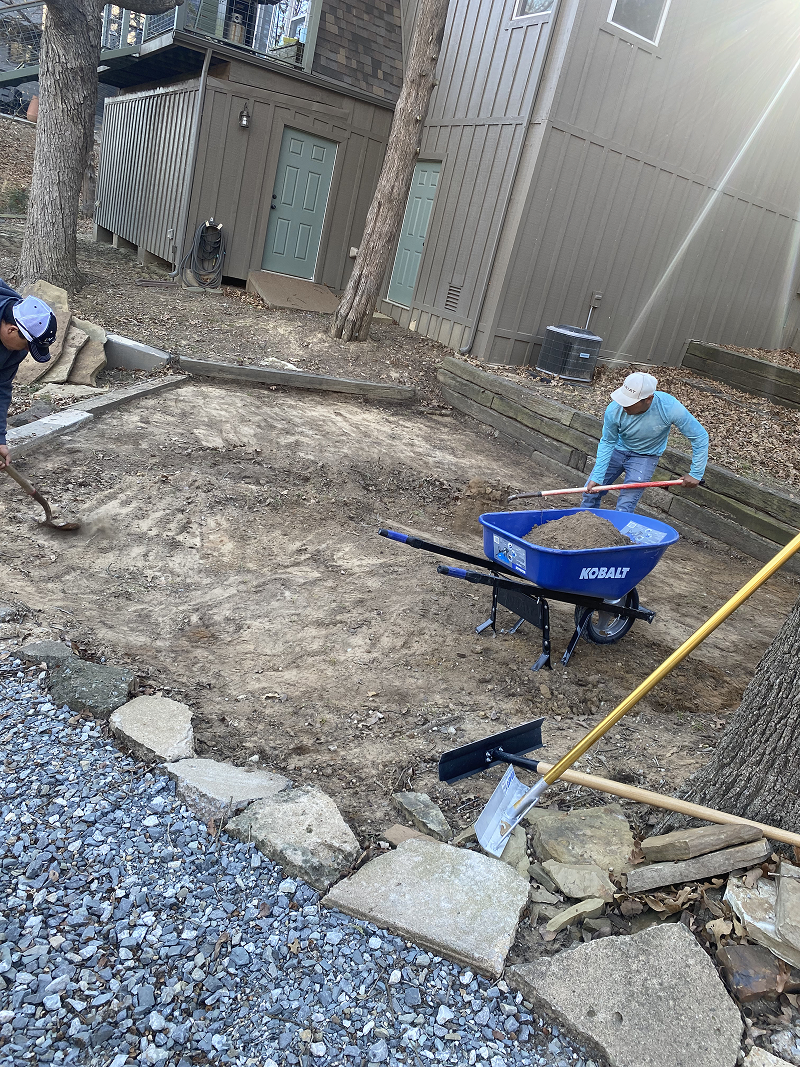 Two men working with shovels, preparing dirt ground near a house with gray siding and green doors, with a blue Kobalt wheelbarrow filled with dirt nearby.