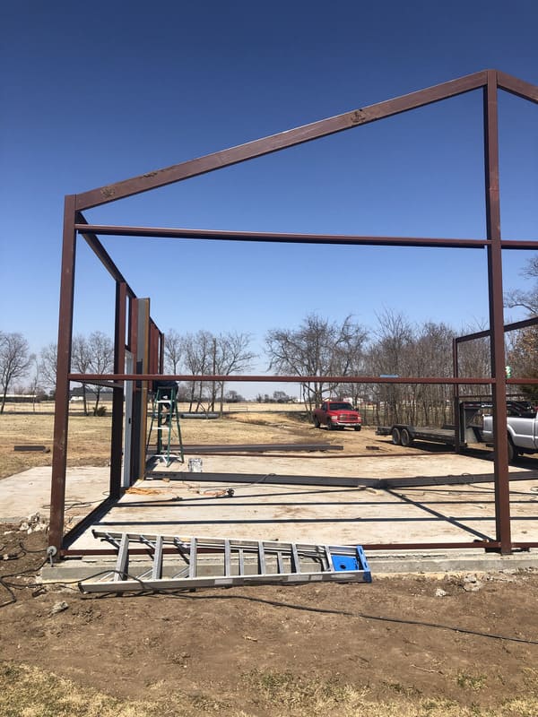 Steel framework of a building under construction on a concrete slab, with a ladder and ladder, set in a rural area with trees and vehicles in the background.