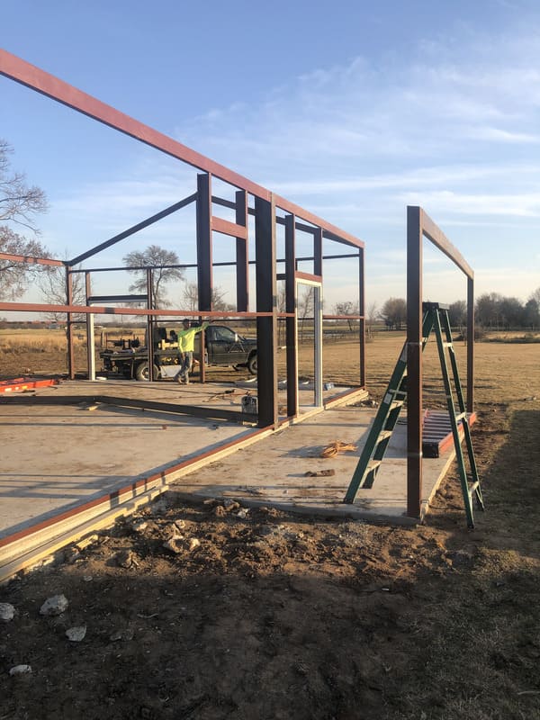Steel frame structure under construction on a concrete foundation in an open field, with a worker and truck in the background and a green ladder nearby.