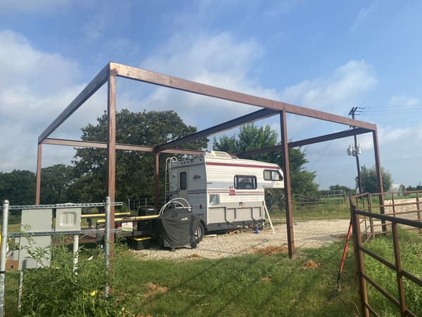Outdoor metal frame structure surrounding a mobile camper trailer parked on gravel with green grass and trees in the background under a partly cloudy sky.