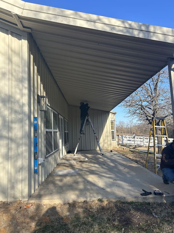 Person standing on a ladder working under a metal roof awning attached to the side of a beige building on a sunny day.