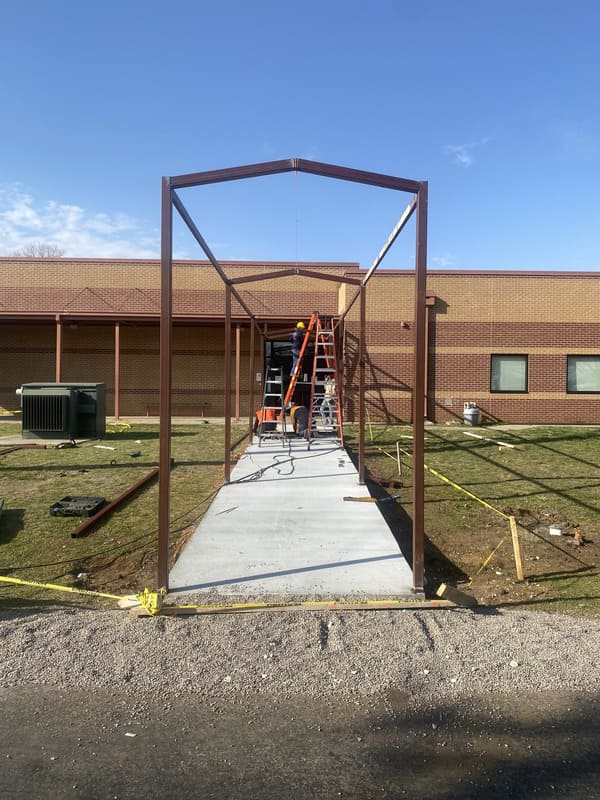 Workers on ladders assembling a metal frame structure over a concrete pathway in front of a brick building under a clear blue sky.