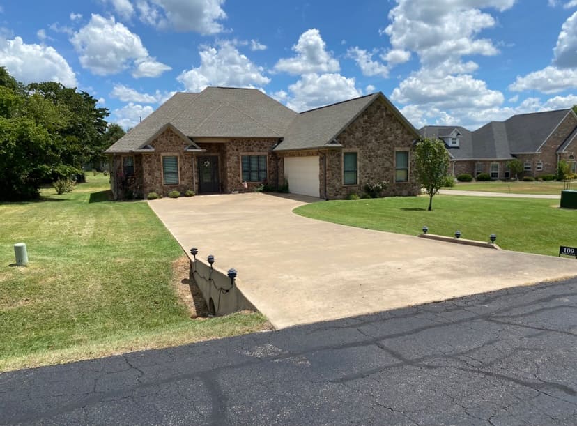 Single-story brick house with a two-car garage, concrete driveway, and lawn under a partly cloudy blue sky.