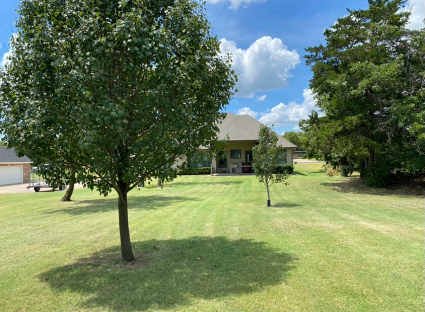 Large grassy yard with several trees leading up to a single-story house with a covered porch under a blue sky with scattered clouds.