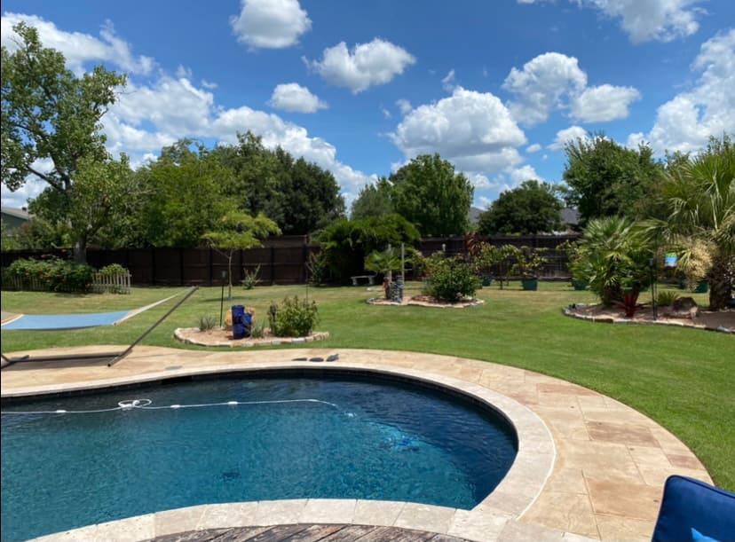 Backyard with a curved swimming pool, surrounded by stone tiles, green lawn, various trees, and a partly cloudy blue sky.