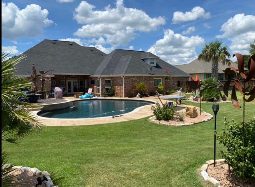 Backyard of a brick house featuring a kidney-shaped swimming pool, patio furniture, and a green lawn under a partly cloudy sky.
