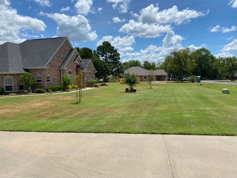 Suburban brick house with a large, well-maintained green lawn under a partly cloudy blue sky.