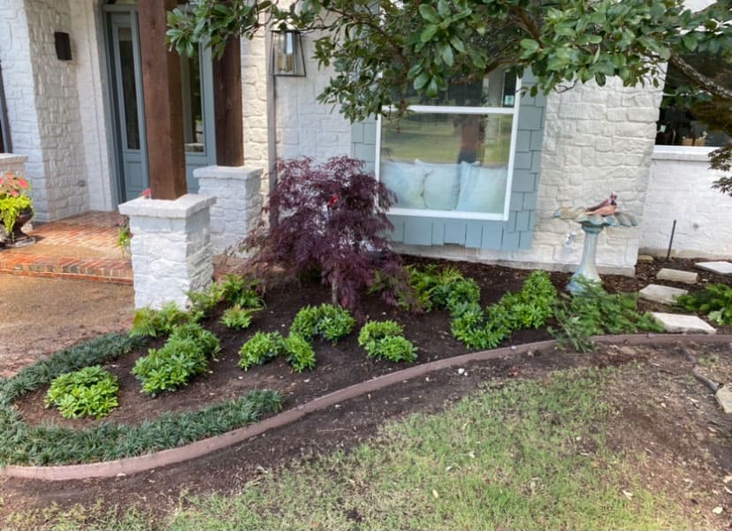 Front yard garden bed with green plants arranged to spell 'GRATEFUL' in front of a house with a white stone exterior and a blue-gray window frame.