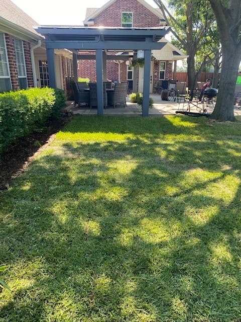Backyard with a green lawn, a pergola covering outdoor dining furniture, and a tree casting shadows on the grass.