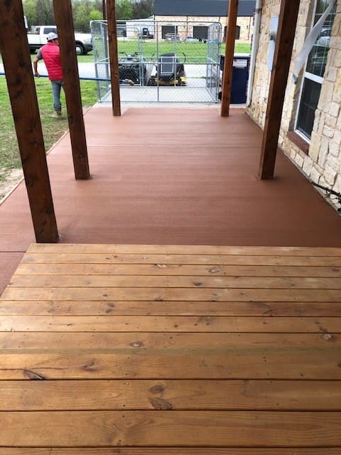 Wooden deck with natural finish leading to a covered porch with brown concrete floor and wooden support posts, view of a fenced backyard and parked vehicles.