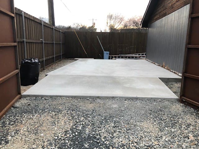 Newly poured concrete slab next to a wooden fence and brown building siding at dusk.